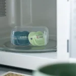 Комплект от 2 бр. ортодонтични залъгалки Tommee Tippee ANY TIME със симетричен силиконов биберон, 6-18м, синя и зелена с декор KIND, в кутия за стерилизиране TT.0274